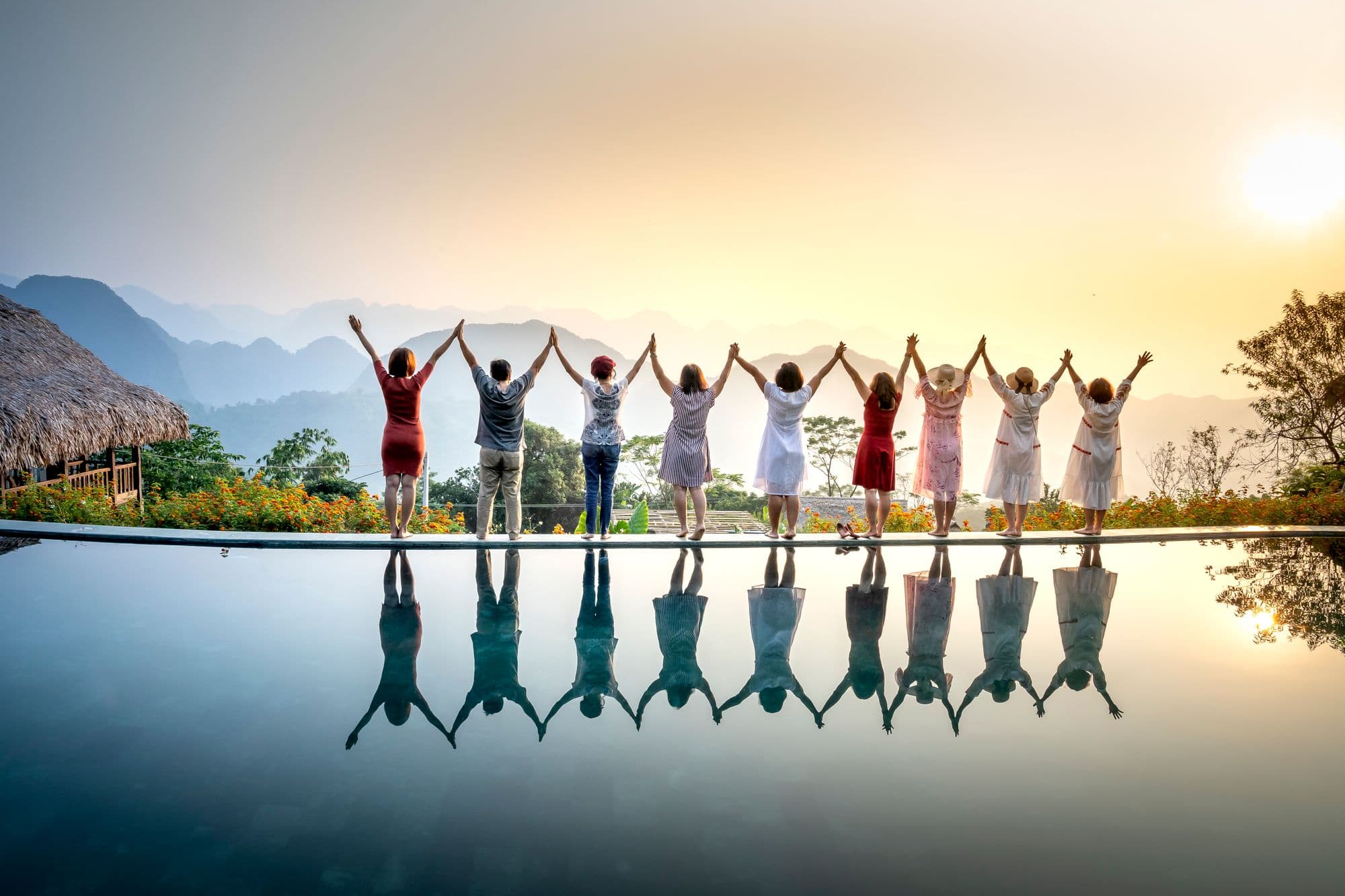 Nine people stand in a row with their backs turned, holding hands with their arms raised, above a still pool at sunrise with mountains behind them.